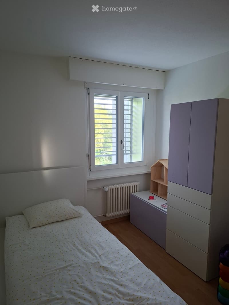 A small, well-lit bedroom with a single bed, a white headboard, a window with blinds, a radiator, a cabinet, and a small wooden shelf.