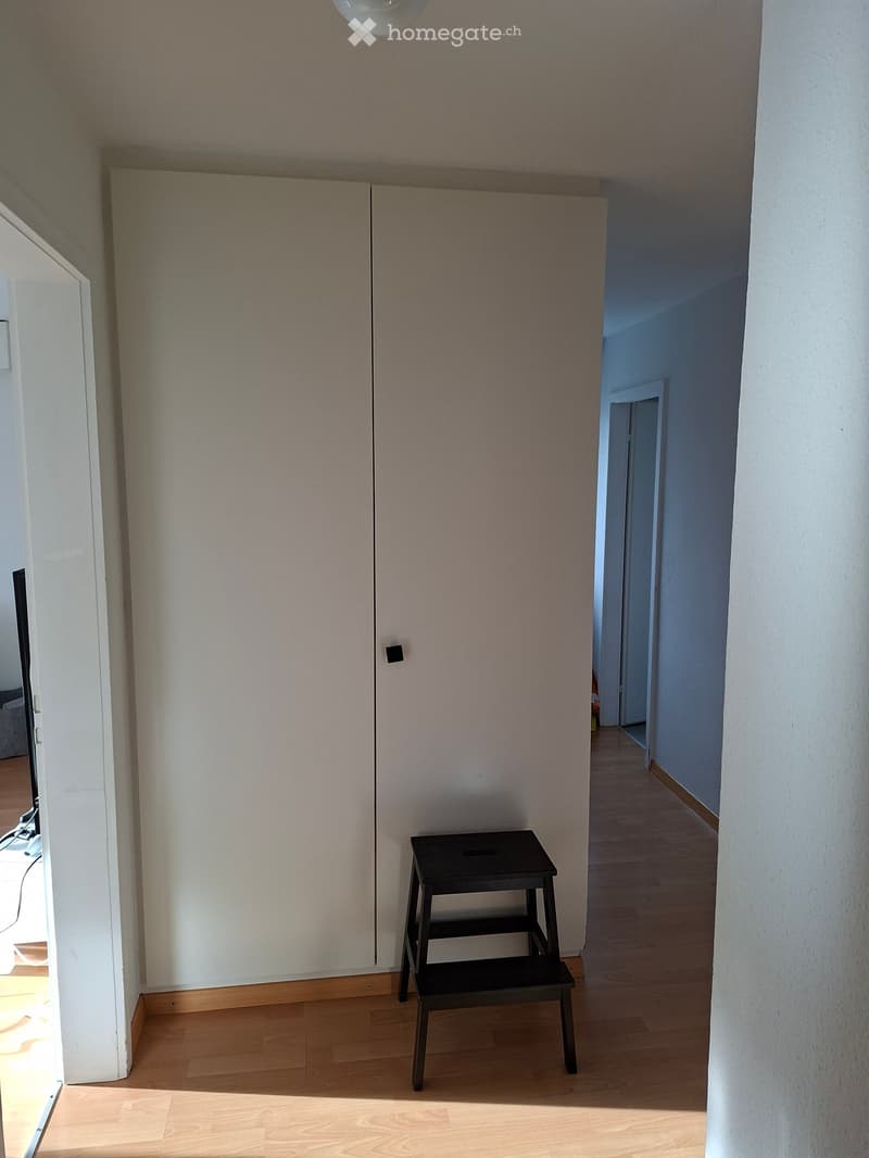 Empty hallway with wooden flooring, white walls, and a small black stool placed in the center.