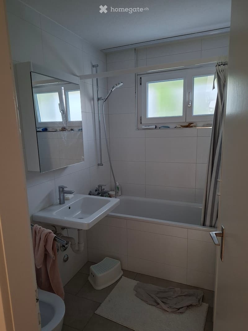 Modern bathroom with white tiles, a sink, bathtub, mirror, and a showerhead. A towel hangs on a rack, and a small stool is placed near the bathtub.