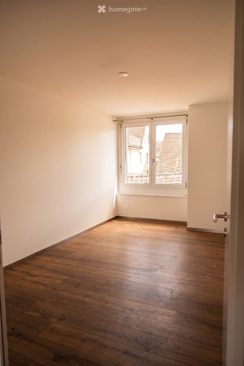 Empty room with wooden flooring, white walls, and a large window with blinds, showing a view of rooftops outside.