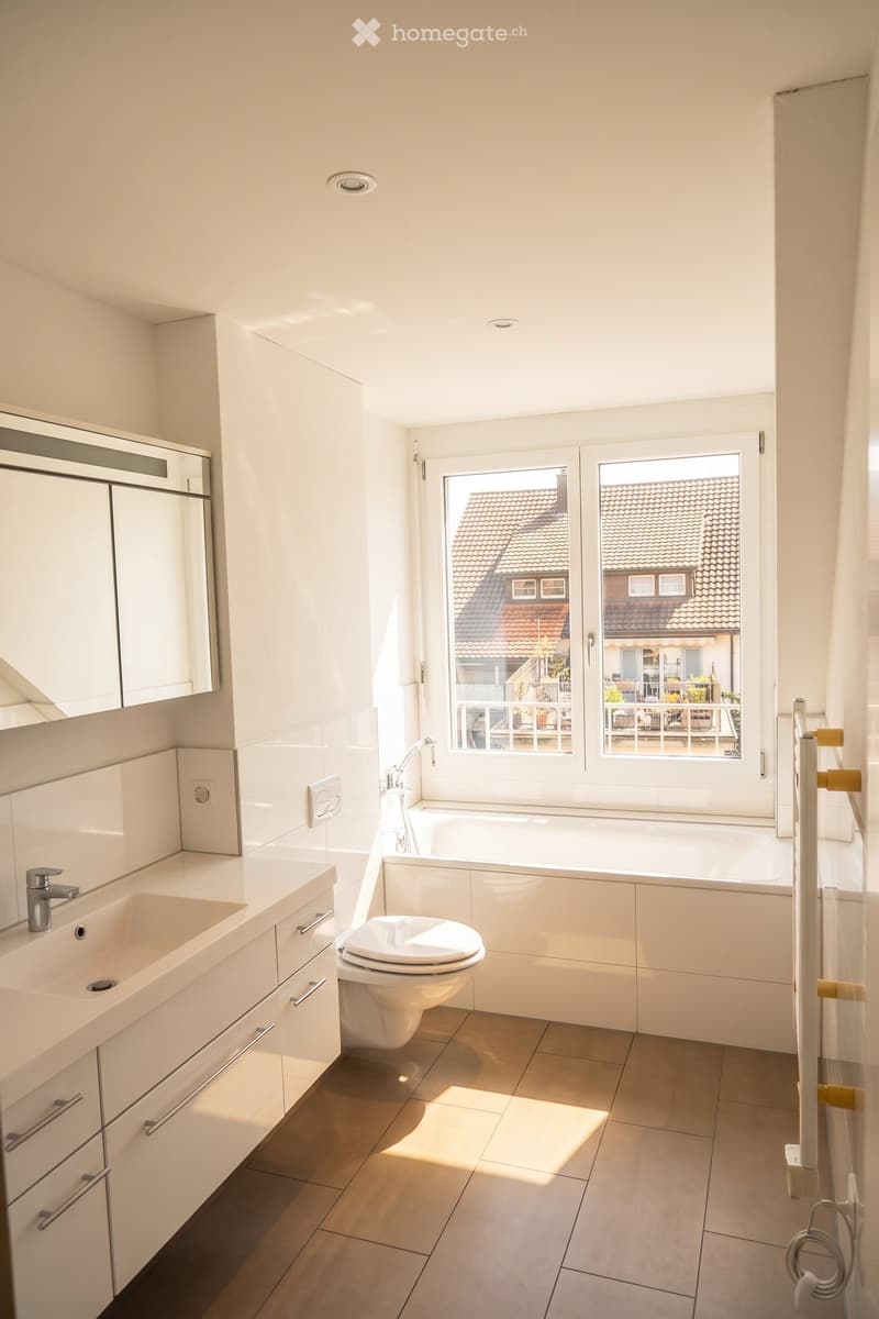 Modern bathroom with white tiles, a large window offering a view of neighboring rooftops, a sink with a modern faucet, a toilet, a bathtub, and a mirrored cabinet.