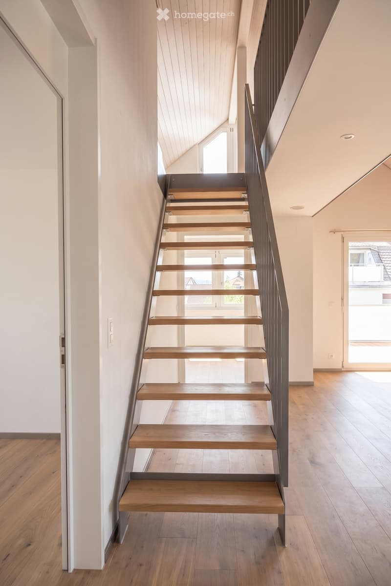 Modern hallway with wooden staircase, white walls, and wooden flooring. Natural light from windows illuminates the space.