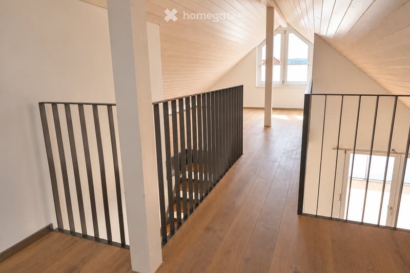 Spacious hallway with wooden flooring, white walls, and modern metal railings leading to a room with large windows and slanted ceilings.
