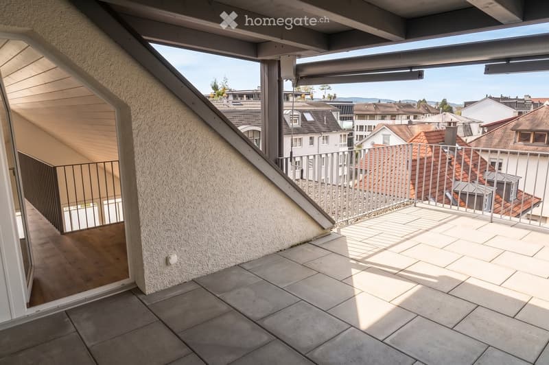 Balcony with tiled floor, metal railing, and a view of neighboring rooftops and a clear sky.