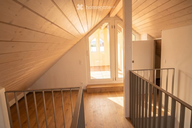 Bright, wooden-floored hallway with slanted wooden ceiling, metal railing, and large window leading to a sunlit area.