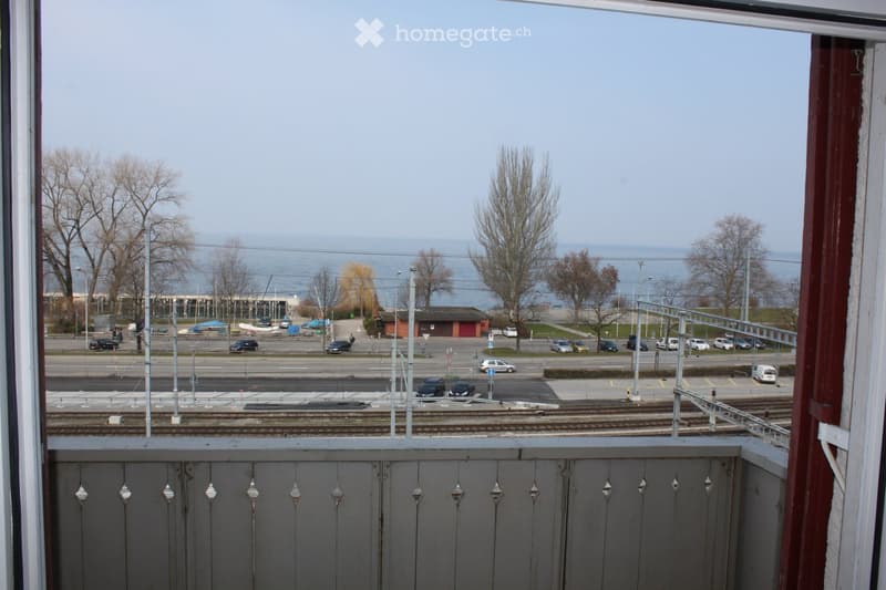 Balcony overlooking a railway track, road, and waterfront with trees and distant water.