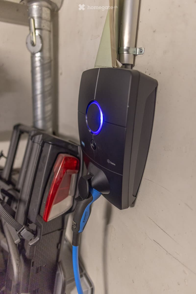 A black electric vehicle charging station mounted on a wall with a blue indicator light and a connected charging cable.