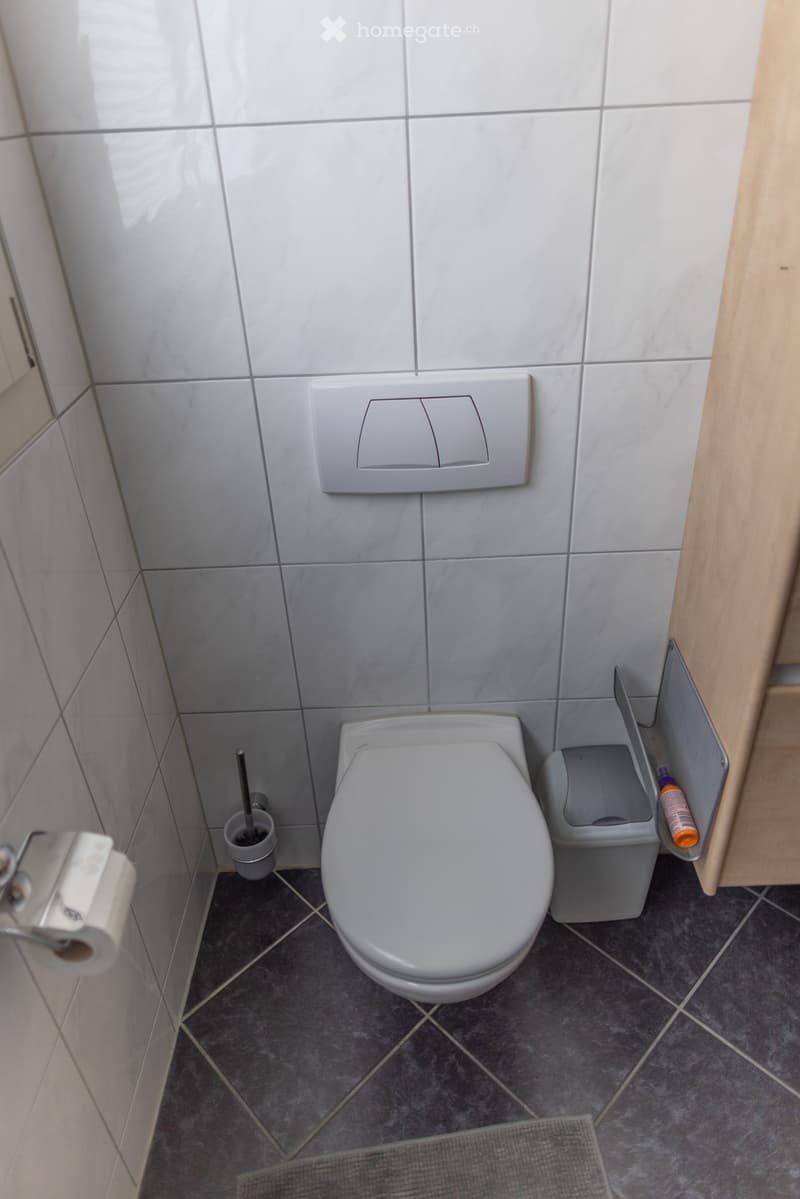 Modern bathroom with white tiled walls, grey tiled floor, toilet, toilet paper holder, toilet brush holder, and trash bin.