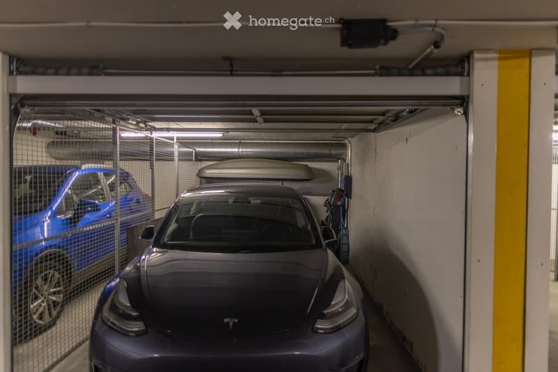 Underground parking area with two cars, one blue and one gray, parked in designated spots. The space features concrete walls, metal railings, and overhead lighting.