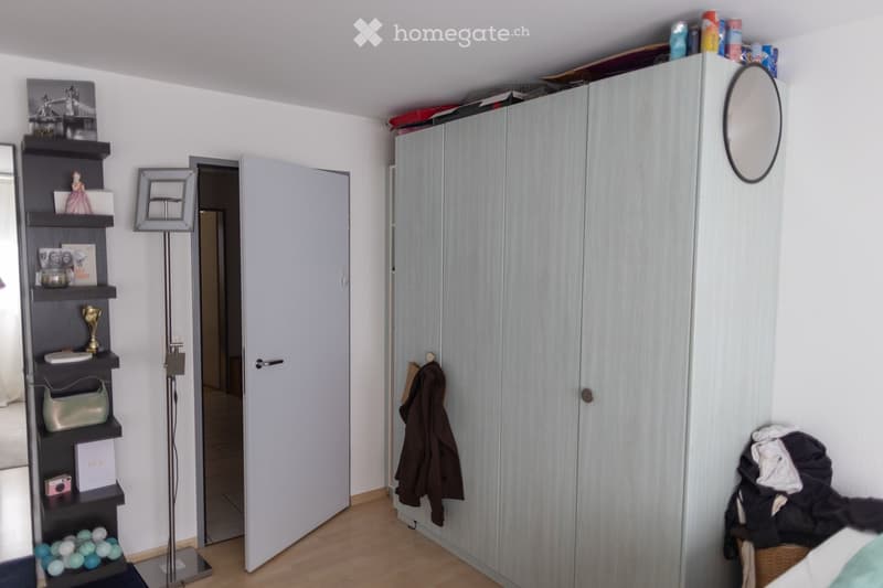 A well-lit bedroom with a large wardrobe, a round mirror, a tall floor lamp, a shelf with decorative items, and a coat hanging on the wardrobe door.