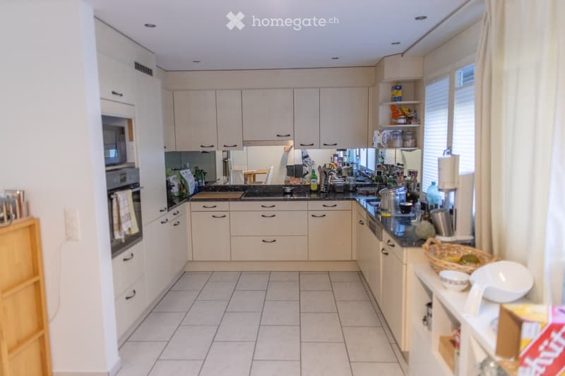 Bright kitchen with white cabinets, tiled floor, and various appliances including a sink, oven, and fridge. Countertop has a blender, cups, and a basket with fruit.