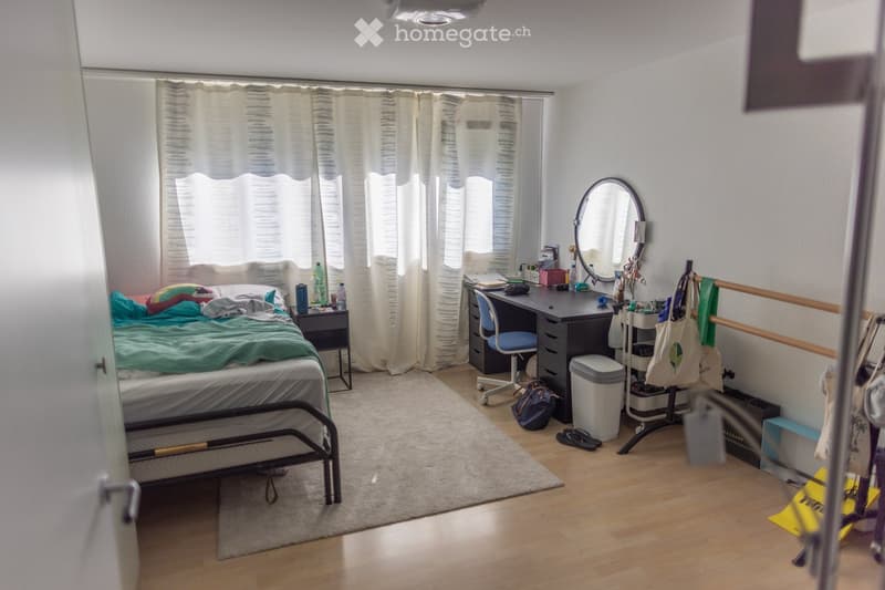 A well-lit bedroom with a bed, desk, chair, mirror, nightstand, and various personal items. The room features wooden flooring and large windows with curtains.