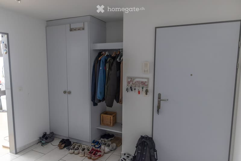 A clean, modern hallway with a built-in wardrobe, coat rack, and several pairs of shoes neatly arranged on the floor. A closed door is visible on the right side.