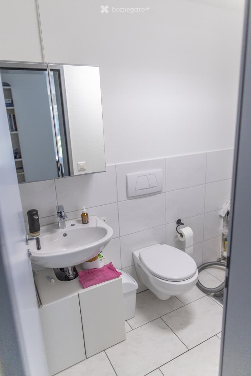 Modern bathroom with white tiles, a sink, toilet, mirror, and a small cabinet. A pink towel is placed on the cabinet.