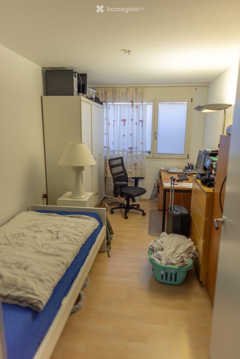 A small, furnished bedroom with a single bed, desk, chair, wardrobe, and a window with curtains. The room includes a laptop on the desk, a suitcase, and a laundry basket.