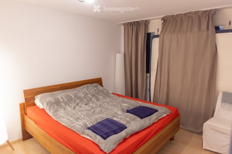A small bedroom with a wooden bed, orange mattress, grey duvet, and two blue pillows. A white chair is positioned near the window with brown curtains.