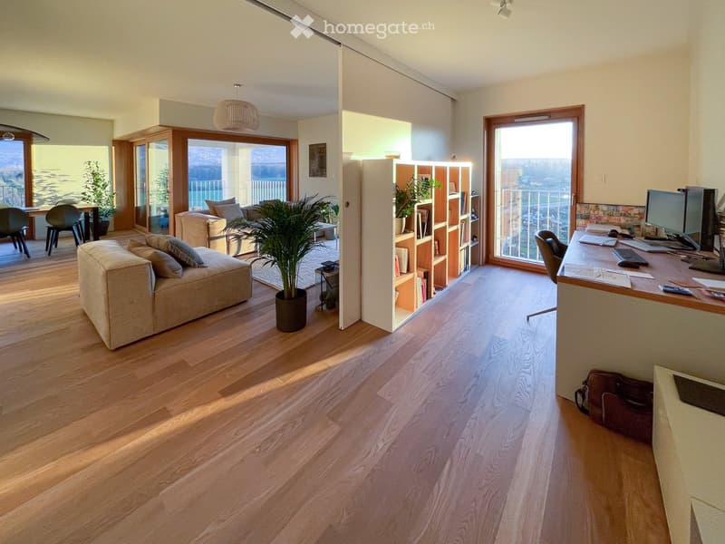 Open-plan living and working space with wooden flooring, large windows offering a scenic view, a sofa, chairs, a dining table, a bookshelf, a desk with a computer, and potted plants.