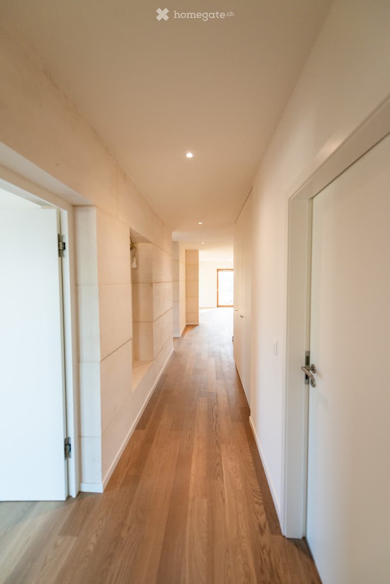 A long, narrow hallway with light wooden flooring, white walls, and recessed ceiling lighting. Doors line both sides, with a small alcove visible on the left.