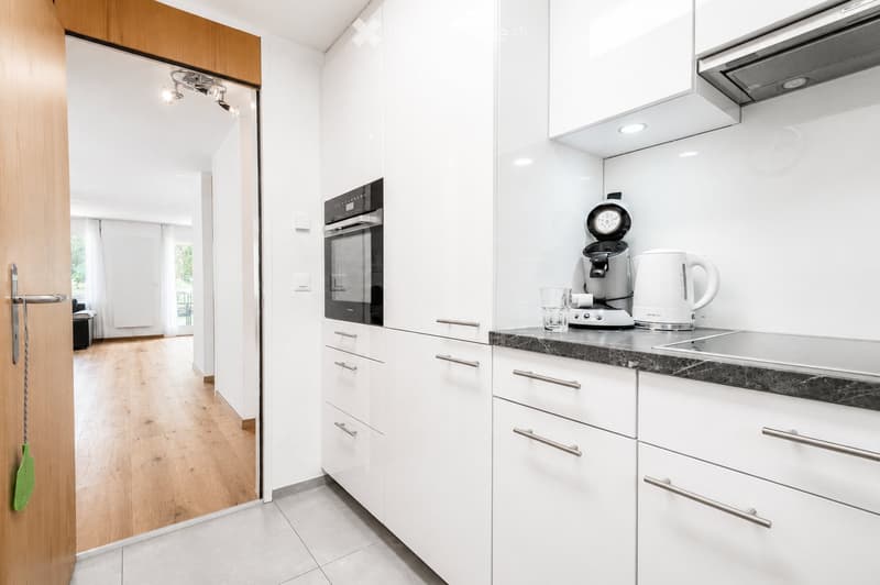 Modern kitchen with white cabinets, stainless steel appliances, and a black countertop. Coffee maker, kettle, and cup are on the counter. An open doorway leads to a living area with wooden flooring.
