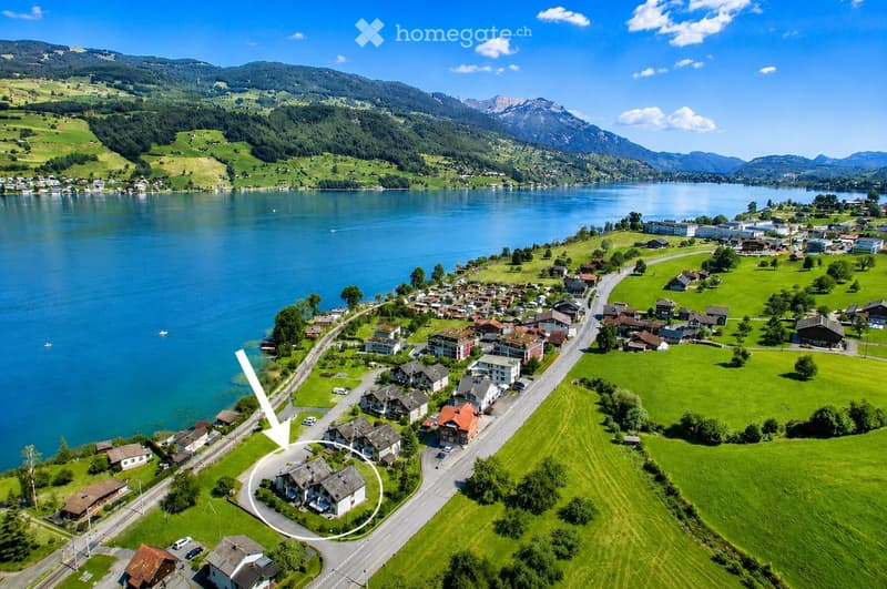 Aerial view of a small village nestled by a lake, surrounded by green hills and mountains. The village features numerous houses with varied architectural styles, roads, and a calm lake reflecting the surrounding landscape.