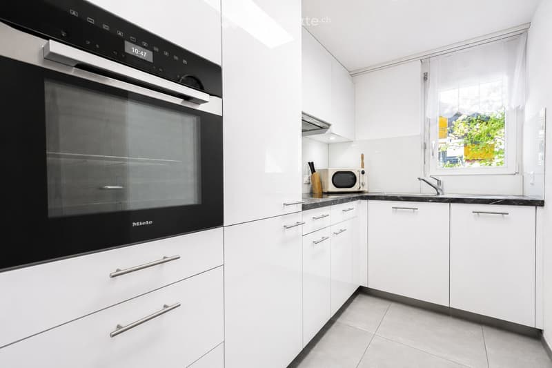 Modern kitchen with white cabinets, stainless steel handles, a built-in oven, microwave, and a sink with a faucet. The countertops are black marble, and there is a window with a view of greenery.