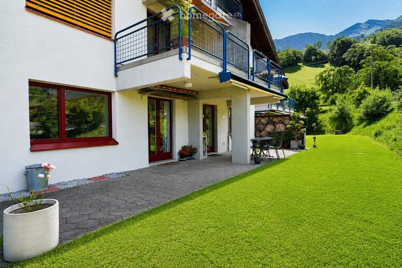 A well-maintained garden area with a green lawn, patio seating, and a view of mountains in the background. The house features a balcony with railings and a stone wall with stacked logs.