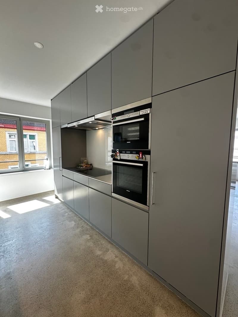 Modern kitchen with grey cabinets, built-in oven, microwave, and a sleek countertop. Natural light from a window illuminates the space.