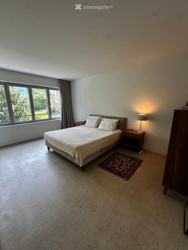 Spacious bedroom with a large bed, nightstand, lamp, and a small rug. Large windows with curtains allow natural light to fill the room.