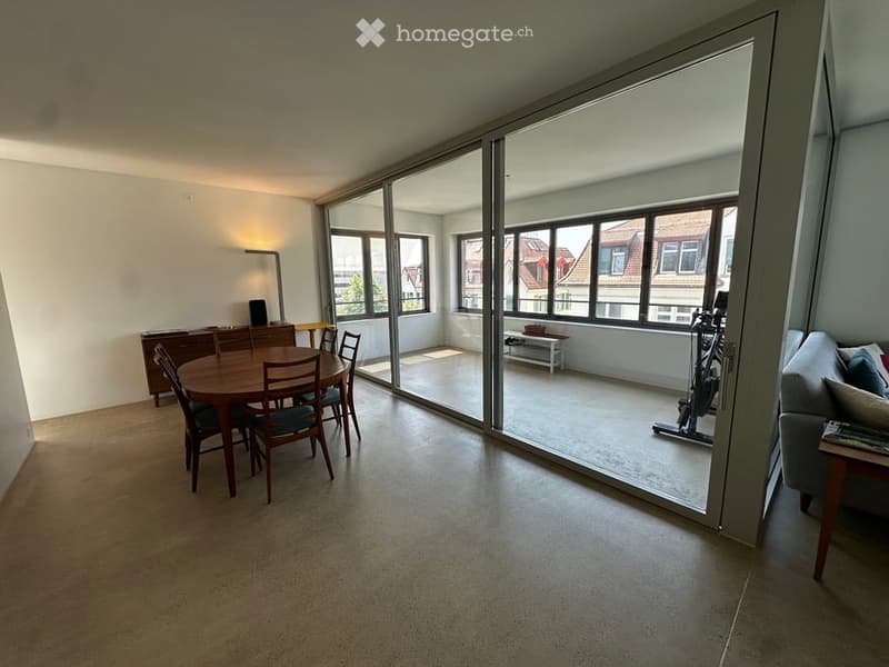 Bright, open-plan living and dining area with large windows and a view of the city. Features a grey sofa, wooden dining table with chairs, a cabinet, and a floor lamp.