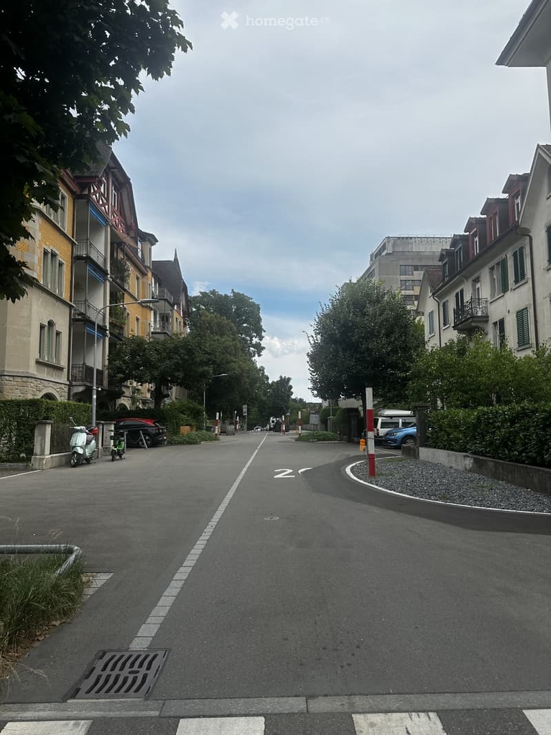 Residential street lined with multi-story buildings, featuring balconies, windows, and parked vehicles including a scooter and a car. The road is marked with white lines and numbers, and there are trees and greenery along the sidewalk.