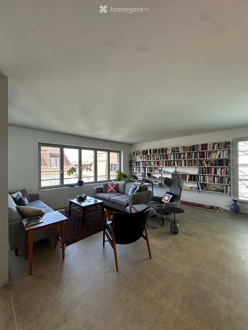 Bright living room with large windows, grey sofa, coffee table, bookshelf, and decorative plants.