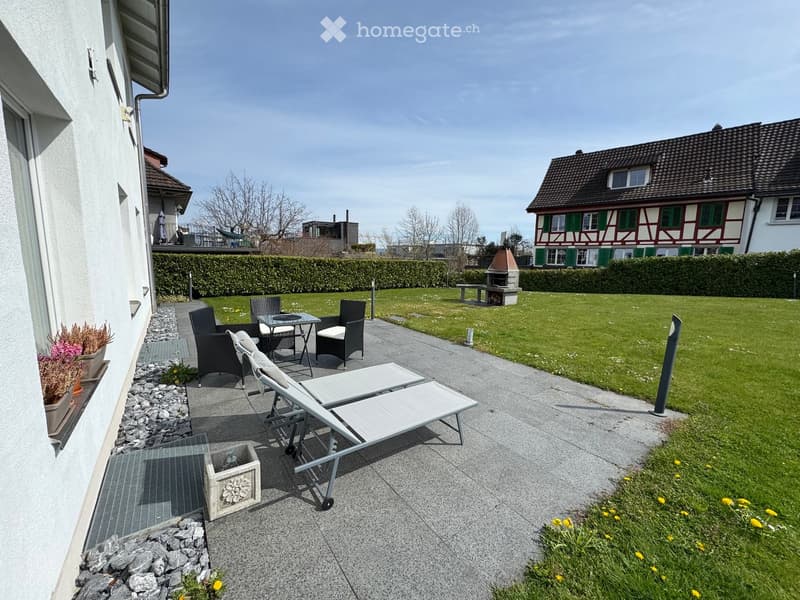 Garden area with a patio, lounge chairs, a small table, and a bench near a barbecue. The area is surrounded by a hedge and a lawn, with a house and additional outdoor furniture visible in the background.