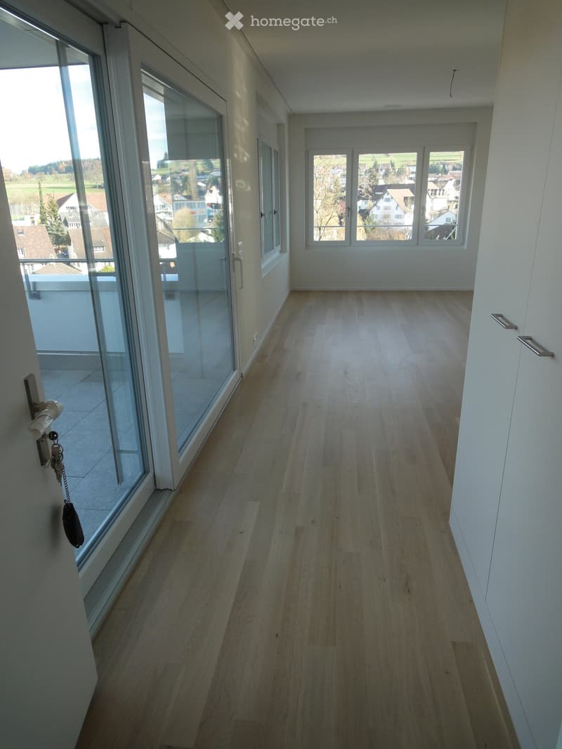 Empty hallway with light wooden flooring, white walls, large windows, and a glass door leading to a balcony.