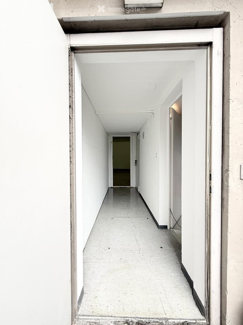 A long, narrow hallway with white walls and a concrete floor, featuring several doors and a visible room at the end.