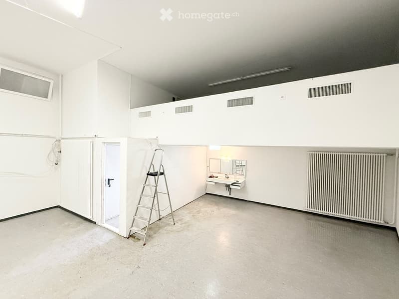 Empty room with white walls, concrete floor, radiator, ladder, and a small bathroom area with sink and mirror.