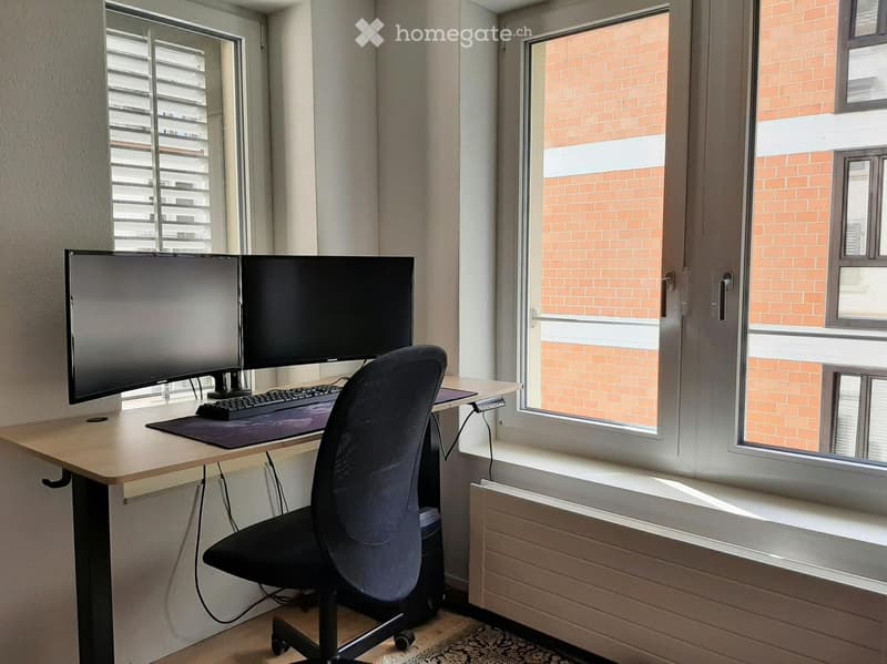 A modern, well-lit room with a desk setup featuring a large monitor, keyboard, and mouse, accompanied by a comfortable office chair. The room has large windows with blinds and a radiator below them.