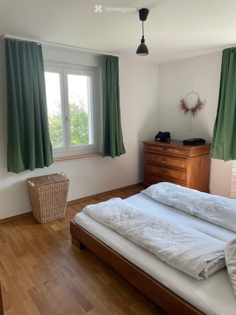Bedroom with wooden floor, two beds, green curtains, wooden dresser, wicker basket, and a lamp