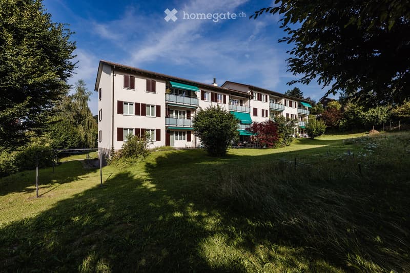 White building with multiple balconies, set in a scenic setting with greenery and trees.