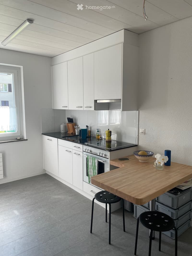 modern kitchen with white cabinets, black countertop, gas stove, oven, white tiles, wooden dining table, two black stools, storage boxes, window with curtains