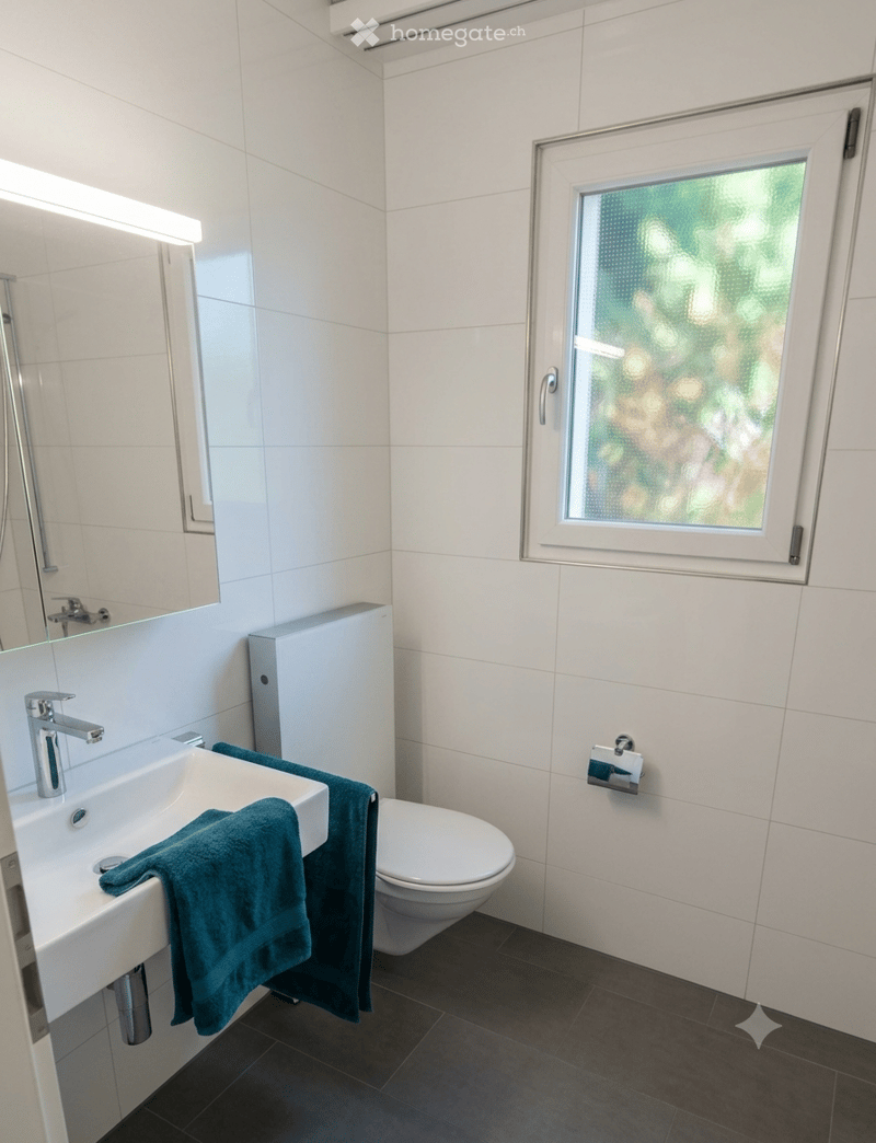 Modern bathroom with white tiles, a sink with a chrome faucet, a toilet, a towel on the sink, and a large mirror above the sink. There is a window with frosted glass on the right side.