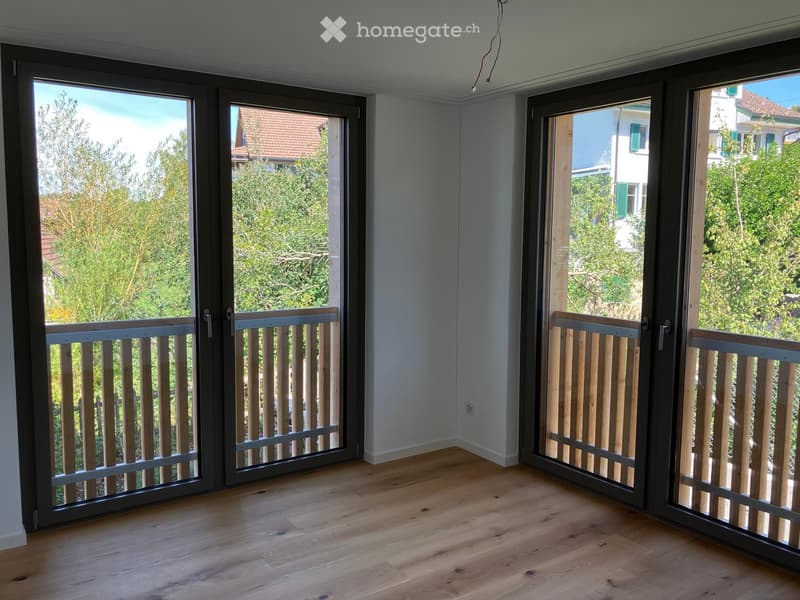 Empty room with wooden flooring and large sliding glass doors leading to a balcony with wooden railings. The view shows greenery and neighboring houses.