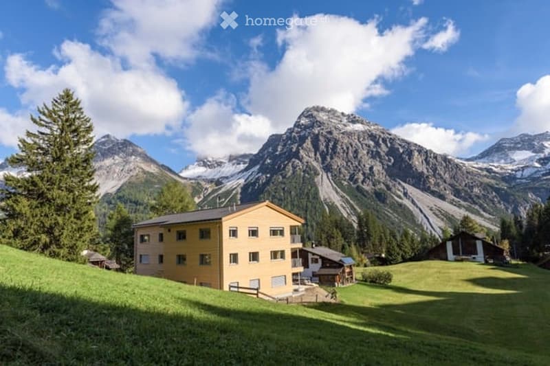 A yellow multi-story building situated on a grassy slope with a panoramic mountain view in the background.