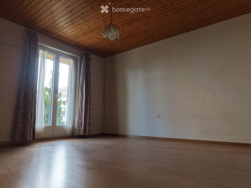 Empty room with wooden flooring, wooden ceiling, large window with curtains, and a hanging light fixture.