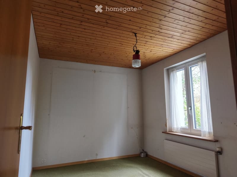 Empty room with wooden ceiling, light-colored walls, green carpet, window with white curtains, radiator, and a hanging light fixture.