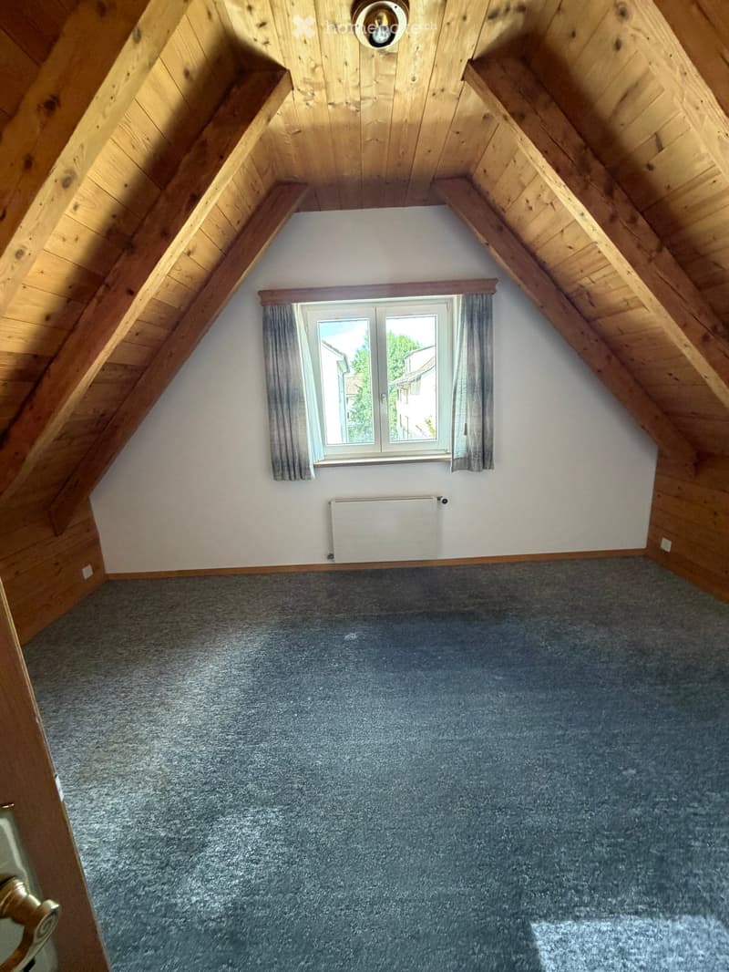 Empty attic room with wooden slanted ceilings, grey carpet, white walls, a window with grey curtains, and a radiator below the window.