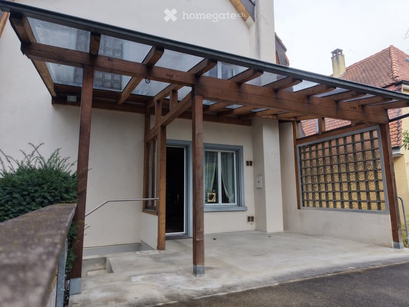 A modern house with a wooden pergola and glass roof, featuring a staircase leading to the entrance, a small garden area, and decorative glass windows.