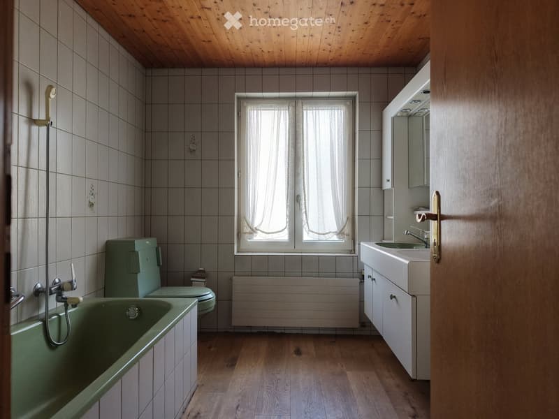 Bathroom with green bathtub, matching toilet, sink with mirror, wooden floor, tiled walls, and a window with sheer curtains.