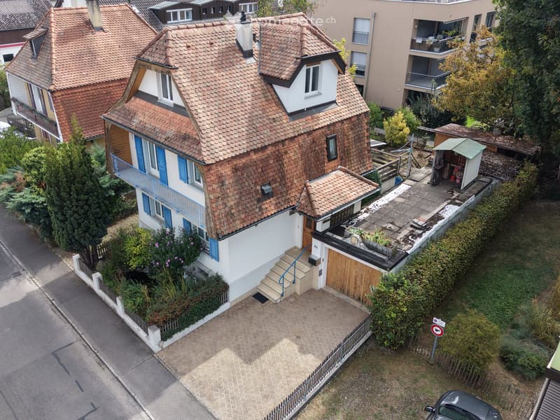 A two-story house with a red-tiled roof, white walls, blue shutters, and a small balcony. The house has a wooden garage and a small garden area with plants and a small shed.