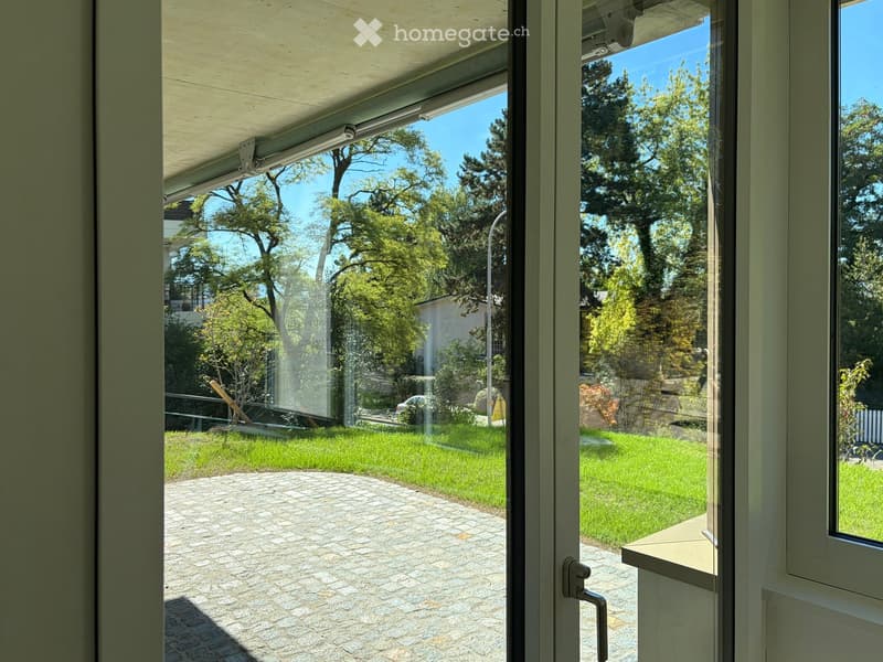 View from inside through large glass doors to a paved patio area with a grassy lawn, trees, and a parked car visible in the background.