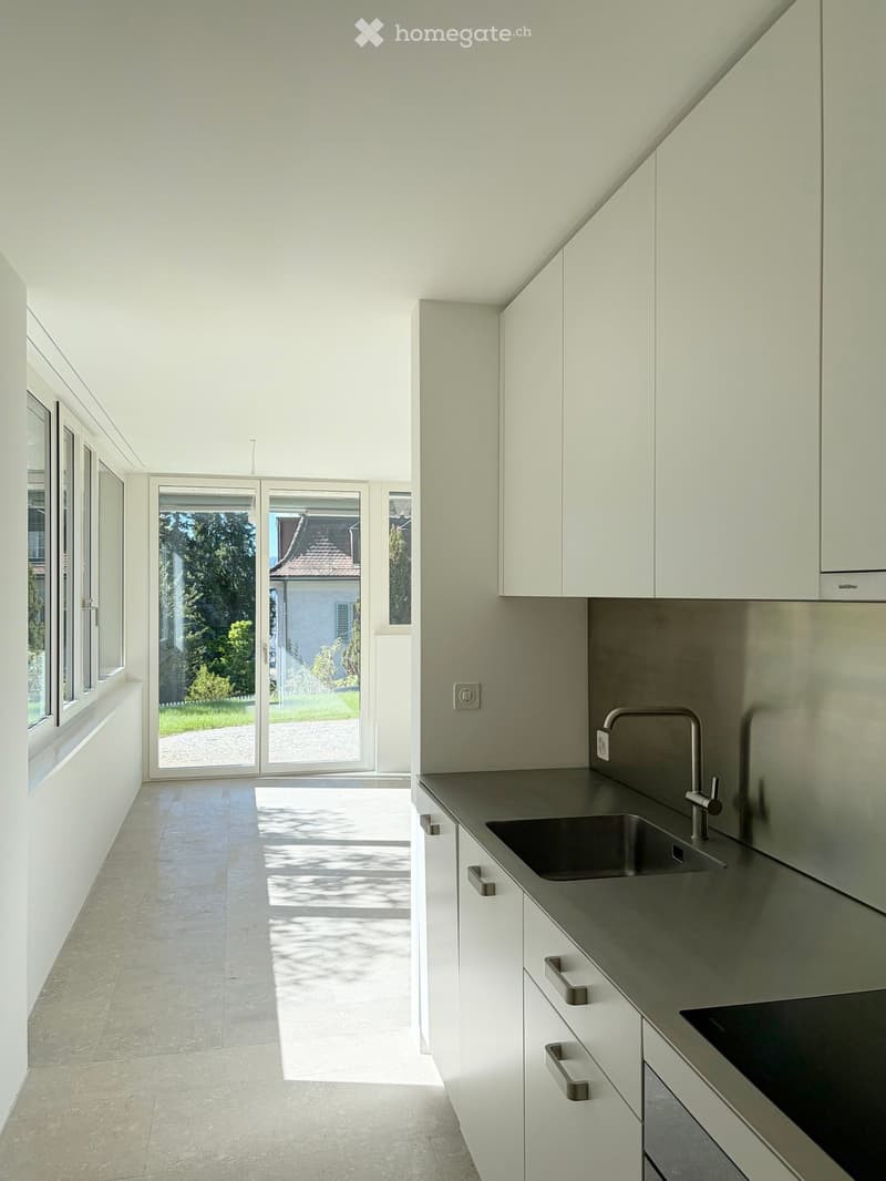 Modern kitchen with white cabinets, stainless steel sink, and large windows overlooking a garden.
